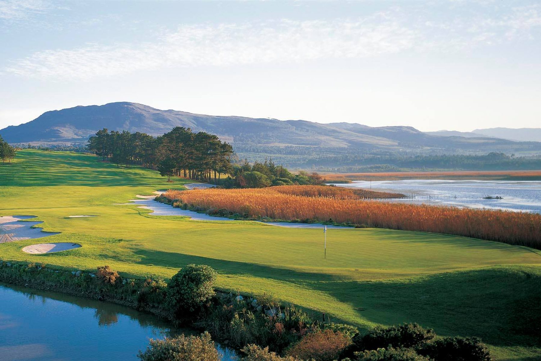 A Gary Player designed golf course at golden hour, with fairways, water and mountains beyond